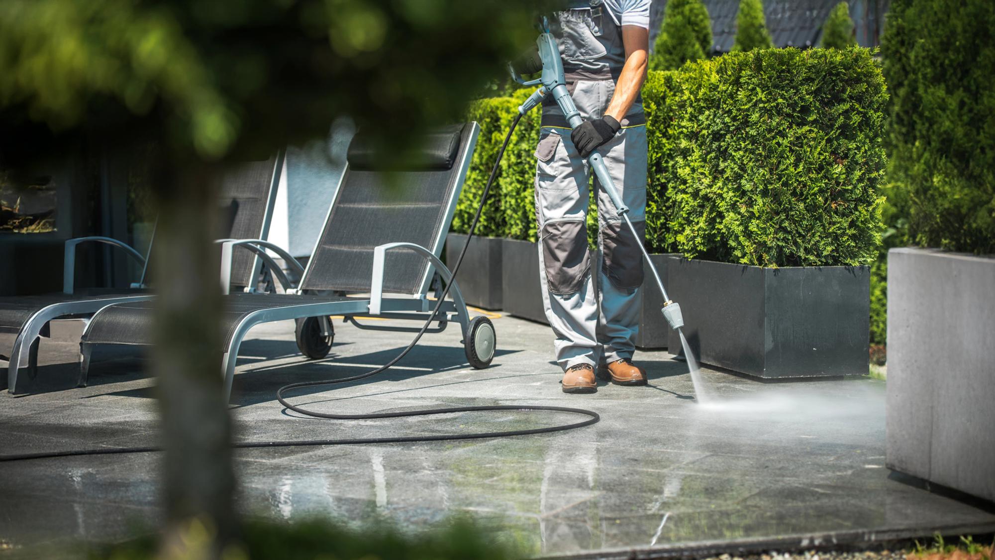 A person in overalls is using a pressure washer to clean a stone patio, with a lounge chair and neatly trimmed hedges visible in the background.